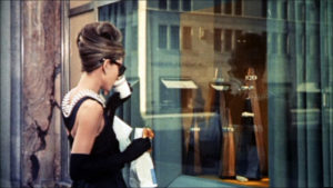 Audrey Hepburn, dressed formally in a black dress with a pearl necklace, looking in the window at Tiffany's jewelry store as she sips on coffee
